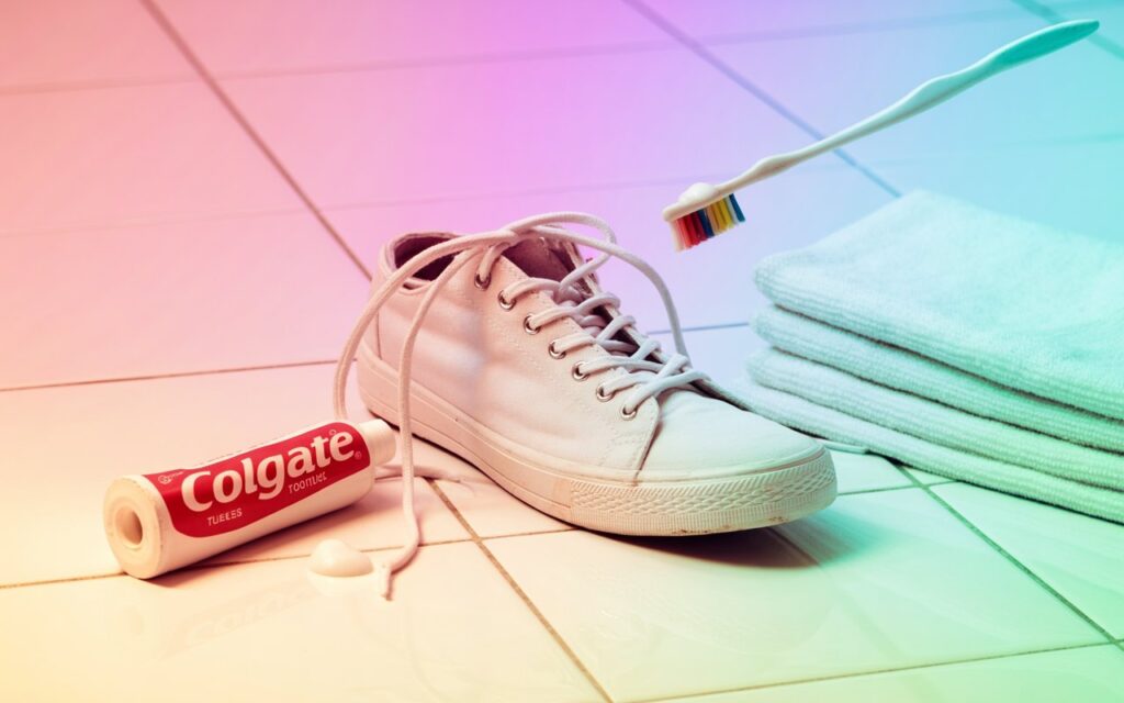 A white sneaker with shoelaces being cleaned using a toothbrush and Colgate toothpaste, placed on a tiled floor with stacked towels in the background.